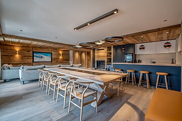 Luxurious, open-plan ski chalet interior with rustic wood walls/ceiling, long dining table, sofa lounge, and modern kitchen bar.