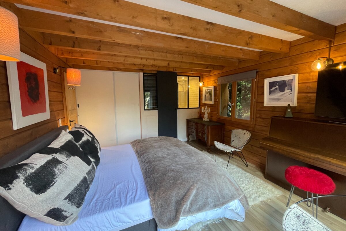A cozy, wood-paneled room with a bed, exposed beams, windows to greenery, rustic furniture, artwork, and a piano. Warm lighting.