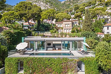 A sleek modern house with a pool and glass walls is built into a lush green hillside, surrounded by trees and other homes under a clear sky.