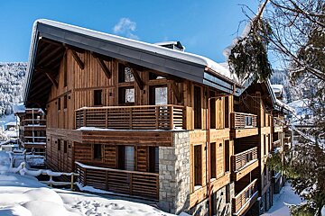 A grand timber chalet with snowy roofs and balconies in a winter mountain setting under a clear blue sky.