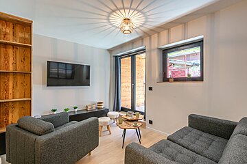 A cozy, modern living room featuring gray sofas, a wall-mounted TV, wooden bookshelf, and hexagonal coffee tables. A glass door and window provide light.