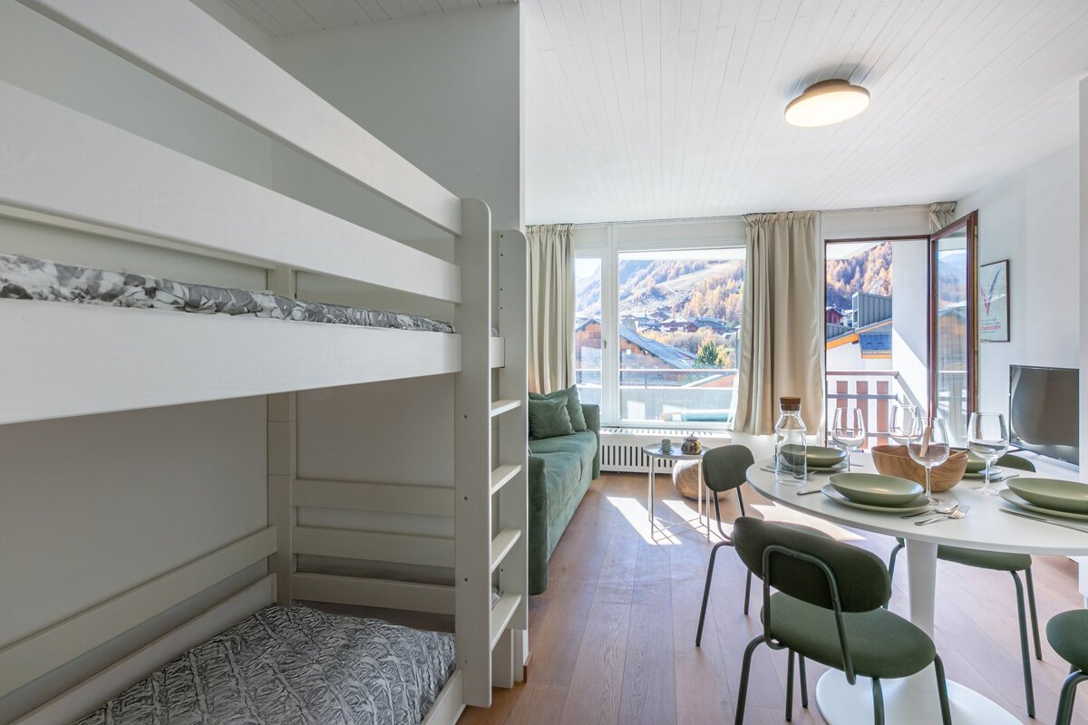 Bright studio apartment featuring white bunk beds, a green sofa, dining table, and a scenic balcony view of autumn mountains & village.