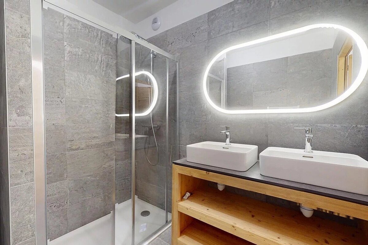 Sleek modern bathroom with a glass shower, double rectangular sinks on a wooden counter, and a backlit oval mirror against gray tile walls.