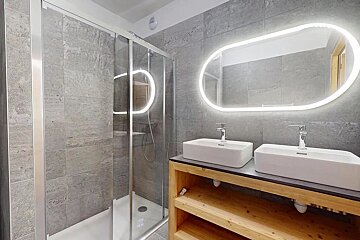 Sleek modern bathroom with a glass shower, double rectangular sinks on a wooden counter, and a backlit oval mirror against gray tile walls.