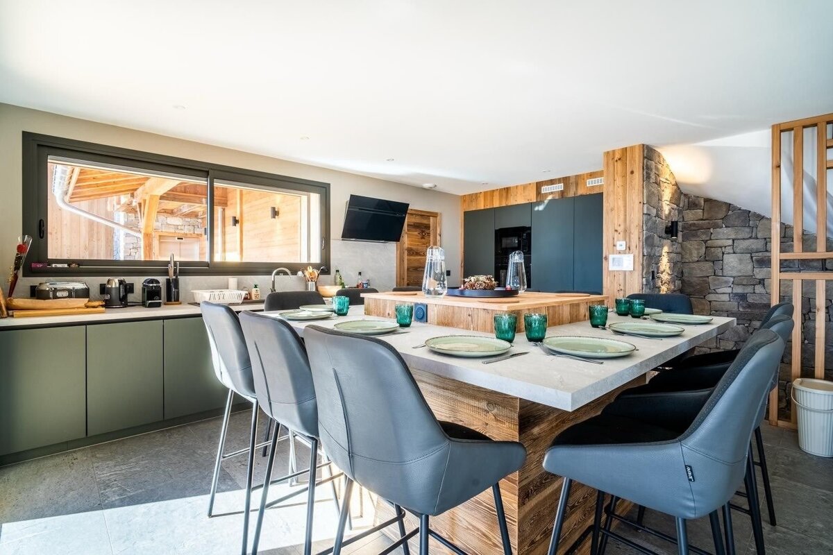 A modern rustic kitchen featuring a large dining island with grey bar stools, set plates, green glasses, and wood and stone accents.