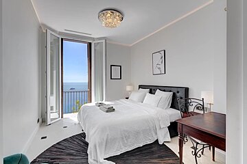 A modern, bright white bedroom with a comfy bed, sparkling chandelier, and a balcony opening to stunning blue sea views.