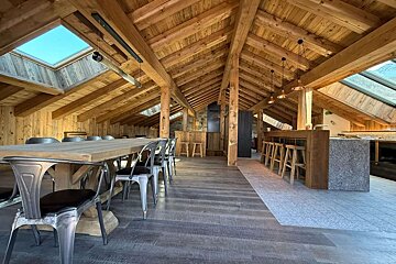 An expansive, modern rustic loft with exposed wooden beams, a large dining area, and a sleek bar/kitchen under skylights.