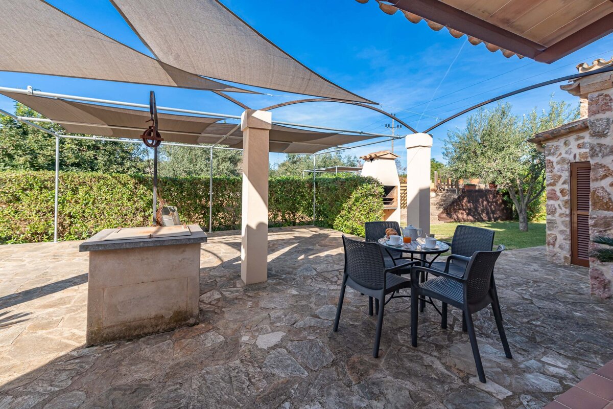 A sunny outdoor patio with shade sails, a table set for breakfast, a well, and a BBQ area, surrounded by lush greenery under a blue sky.