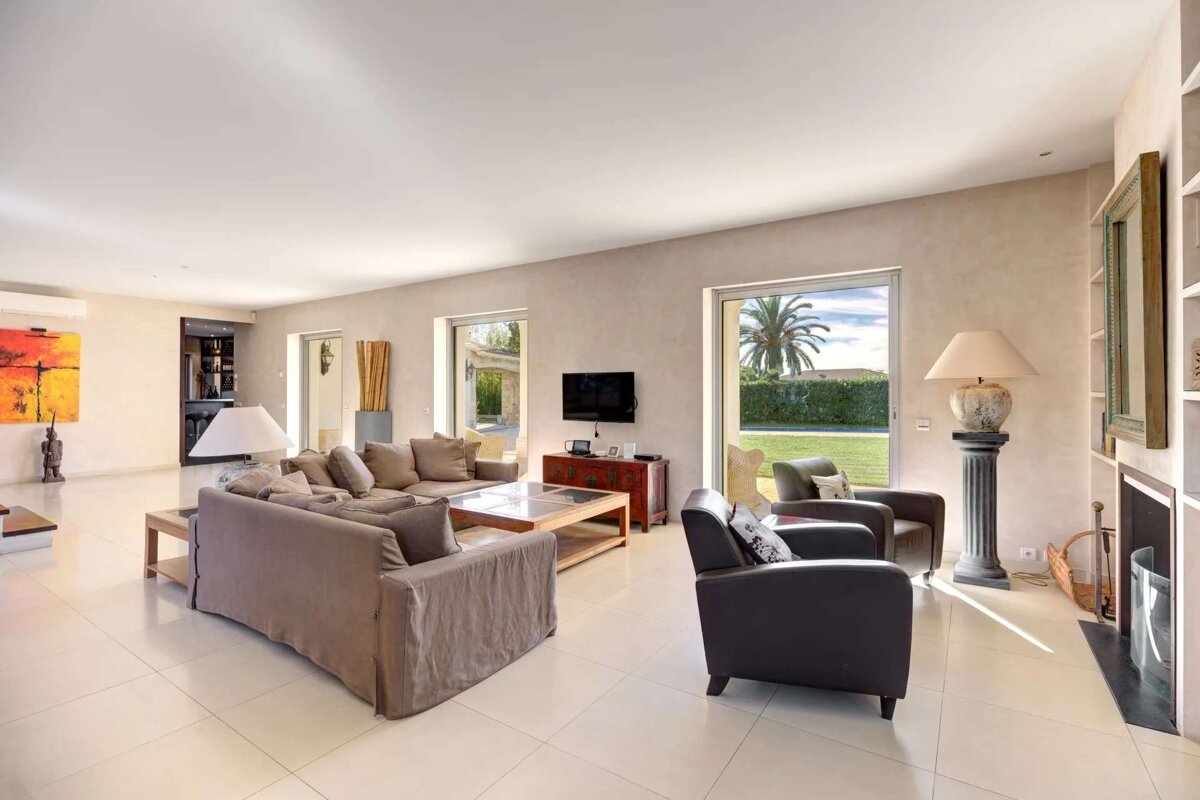 Bright living room with neutral sofas, armchairs, and large windows overlooking a lush green yard with a palm tree. Modern and spacious.