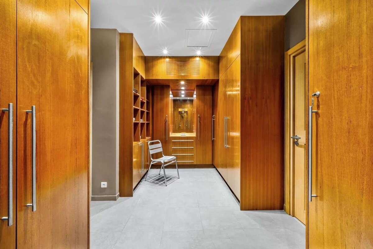 An elegant walk-in closet with sleek wooden wardrobes, a mirrored vanity, storage shelves, a white chair, and grey floor tiles under bright spotlights.