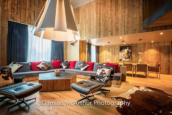 Modern wood-paneled chalet living room with a large sofa, leather chairs, red and cowhide pillows, and a dining area.