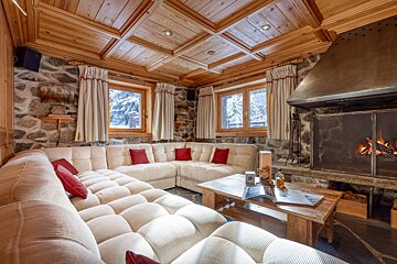 Cozy, rustic lounge with stone walls, wooden ceiling, large cream sofa with red cushions, lit fireplace, and snowy views outside.
