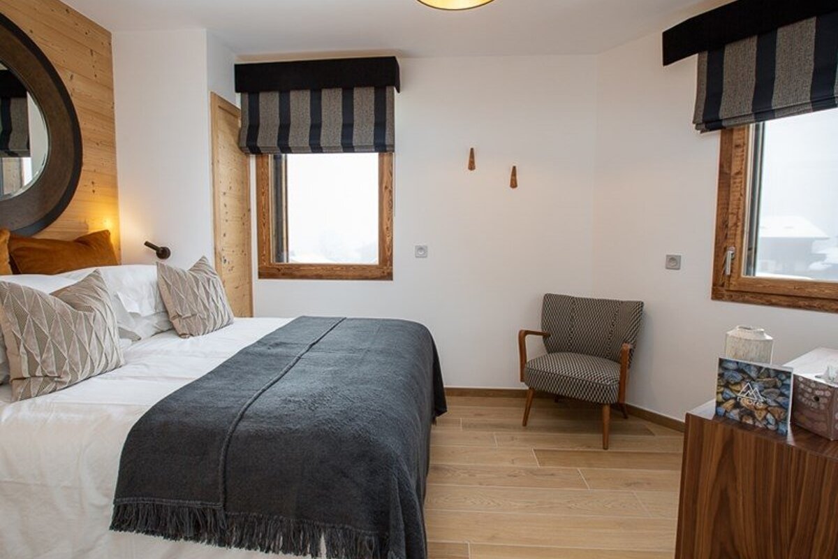 A cozy modern bedroom featuring a bed with a grey throw, striped window shades, a patterned armchair, and light wood flooring.