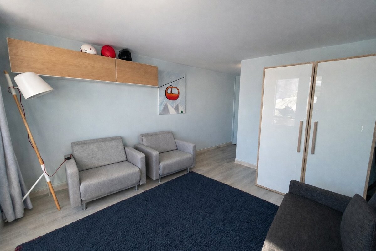 A bright room with light blue walls, a dark blue rug, two grey armchairs, and a floor lamp. Ski helmets on a shelf and gondola art suggest a mountain theme.