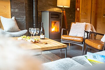 A cozy, rustic living room featuring a lit fireplace, wine and cheese on a coffee table, and comfortable armchairs, evoking a warm and relaxing atmosphere.
