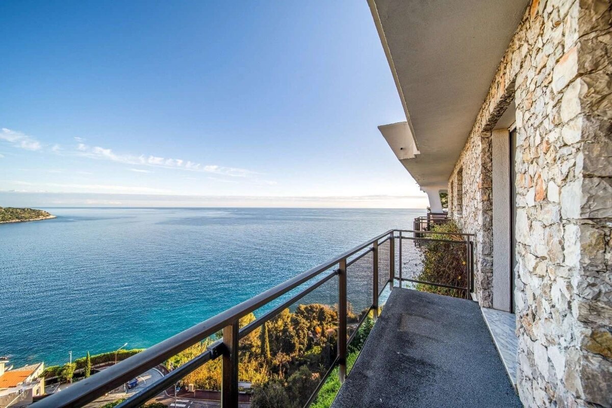 A stone-walled balcony overlooks a vast, clear blue ocean under a bright sky, offering a stunning panoramic coastal view.