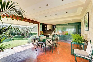 A modern sunroom with green kitchen cabinets, a dining table, hexagonal tile floor, and large windows opening to a lush garden.