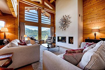 A rustic-modern living room featuring wood walls, exposed beams, large windows overlooking mountains, a sleek fireplace, and comfy sofas.