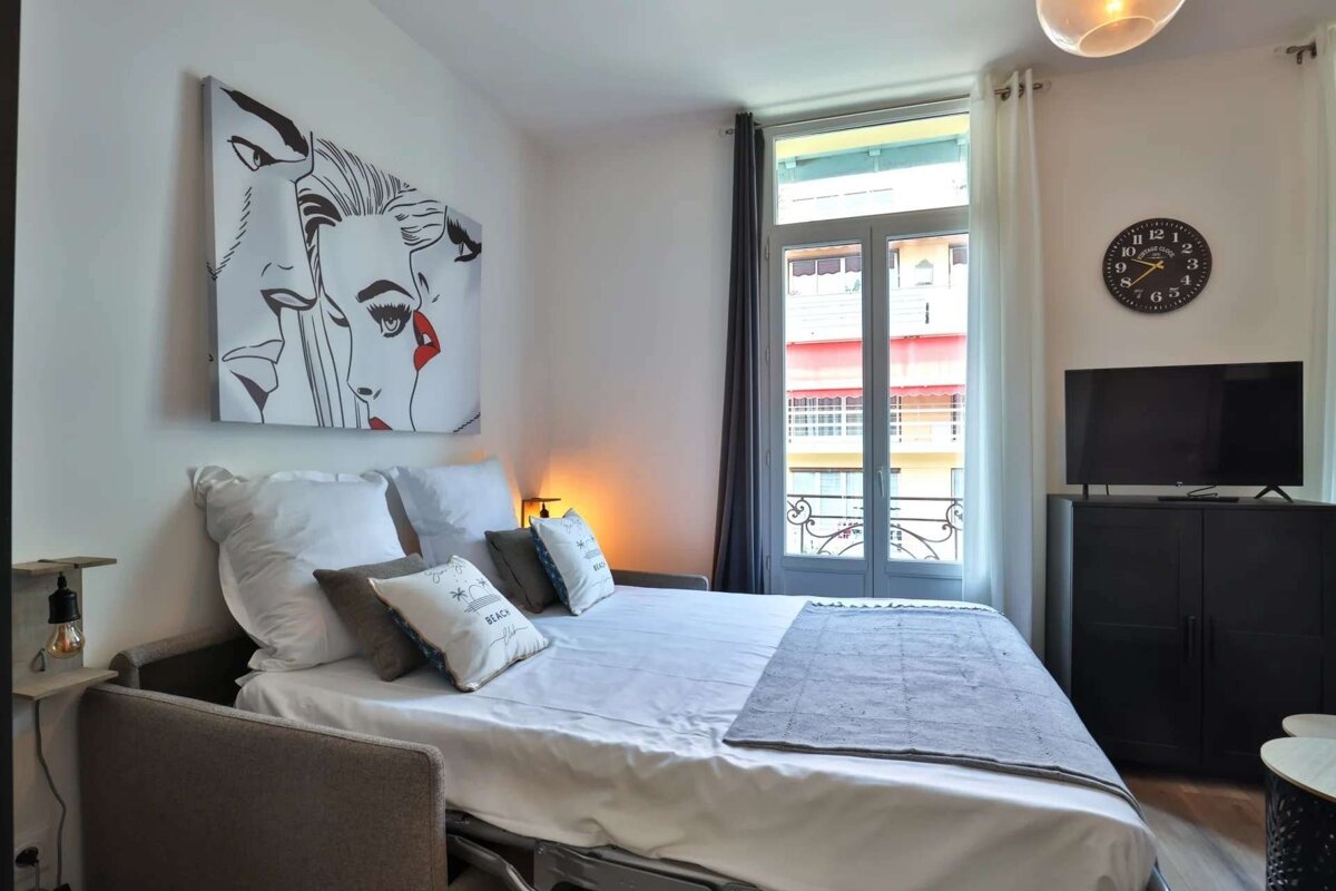 A modern studio apartment with a pull-out sofa bed, pop art, a large window offering an outside view, a TV, and a wall clock.