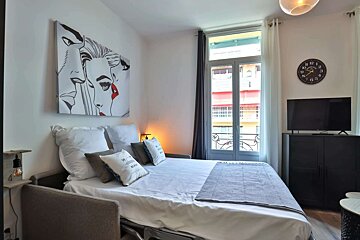 A modern studio apartment features a sofa bed, large pop art painting, balcony door, TV, and black clock on a white wall.