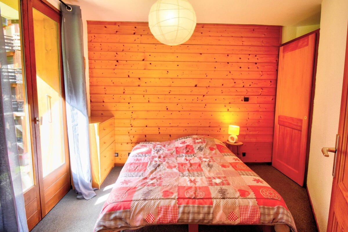 Cozy chalet bedroom featuring wooden paneling, a bed with a patchwork quilt, a large glass door to the left, a ceiling lantern, and a bedside lamp.