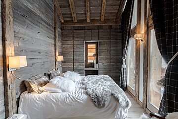 Rustic wooden bedroom featuring a cozy white bed with furry accents, large windows, and dark plaid curtains.