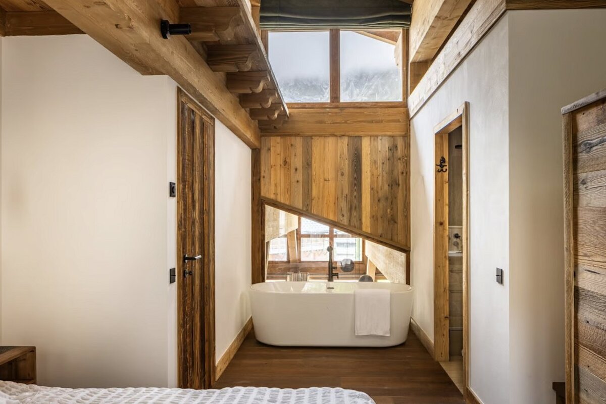 Rustic-modern room with a freestanding tub under a large window, featuring exposed wooden beams, plank walls, and white finishes.