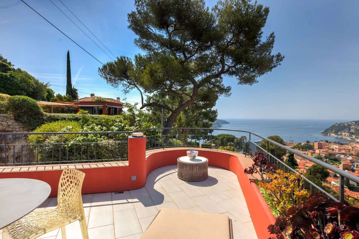 Sunny Mediterranean terrace with red walls, a large pine tree, and a stunning view of a coastal town and the deep blue sea under clear skies.