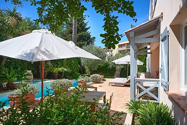 Sunny Mediterranean patio with a swimming pool, white umbrellas, and wooden furniture next to a villa.