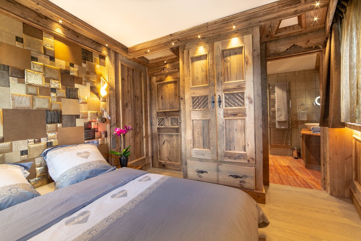 A cozy, rustic bedroom with extensive wood paneling, a bed with heart-patterned linens, and an en-suite bathroom visible.