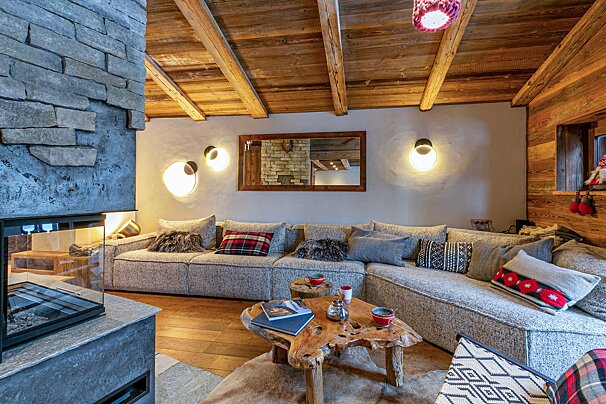 A cozy, rustic-modern chalet living room features a stone fireplace, beamed wooden ceiling, large sectional sofa, and a natural wood coffee table, with warm lighting.