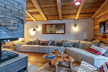 A cozy, rustic-modern chalet living room features a stone fireplace, beamed wooden ceiling, large sectional sofa, and a natural wood coffee table, with warm lighting.