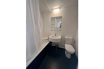 A functional bathroom with a white bathtub and shower curtain, sink, toilet, and mirror. It features white walls and a dark blue floor.