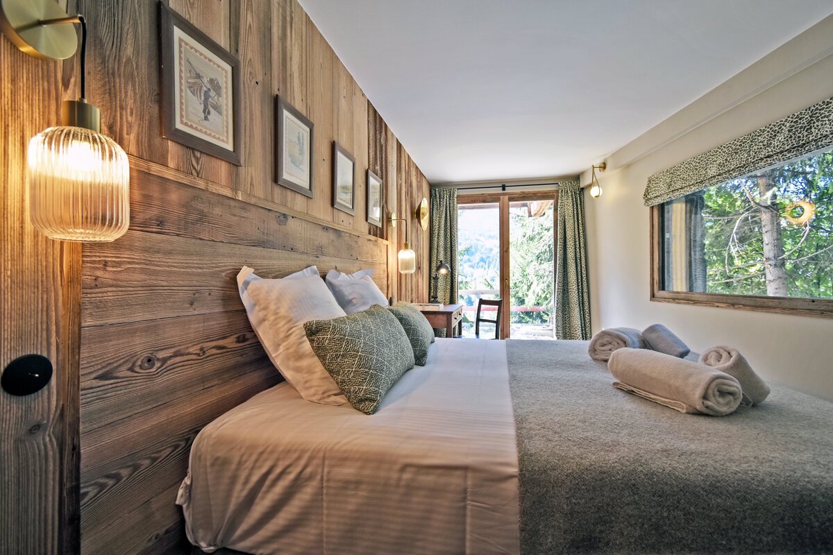 A cozy, modern bedroom with a wood-paneled accent wall, a comfortable bed, and large windows/door offering a scenic view of lush trees.