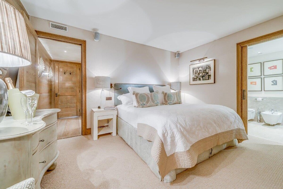 A cozy, neutral-toned bedroom features a large bed with patterned pillows, bedside tables, and lamps. Doors lead to a hallway and a modern bathroom.