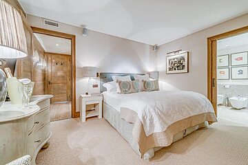 A cozy, neutral-toned bedroom features a large bed with patterned pillows, bedside tables, and lamps. Doors lead to a hallway and a modern bathroom.
