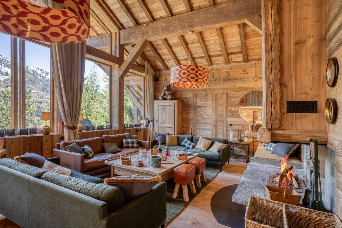 A cozy, rustic chalet living room with wood walls/ceiling, mountain views, a crackling fireplace, and plush sofas creates a warm ambiance.