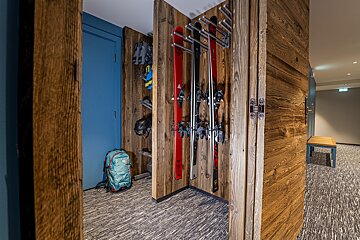 A ski storage area with rustic wooden walls, featuring skis, boots, helmet, and a backpack. A blue door is visible, leading to a hallway.