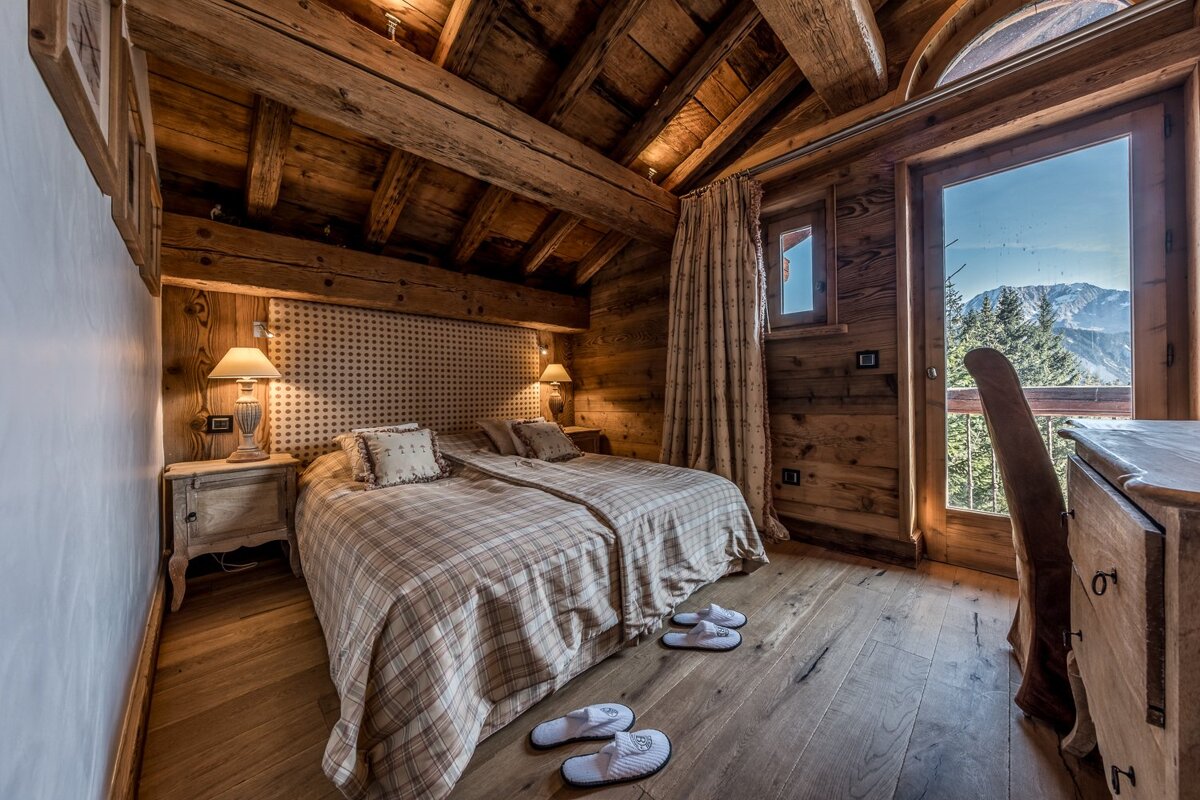 Cozy, rustic wooden bedroom with twin beds, plaid bedding, exposed beams, and a balcony offering a stunning mountain view.