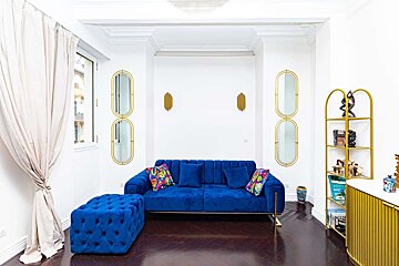 A chic room featuring a royal blue sofa and matching ottoman, gold oval mirrors, sconces, and a shelf, against white walls and dark wood floors.