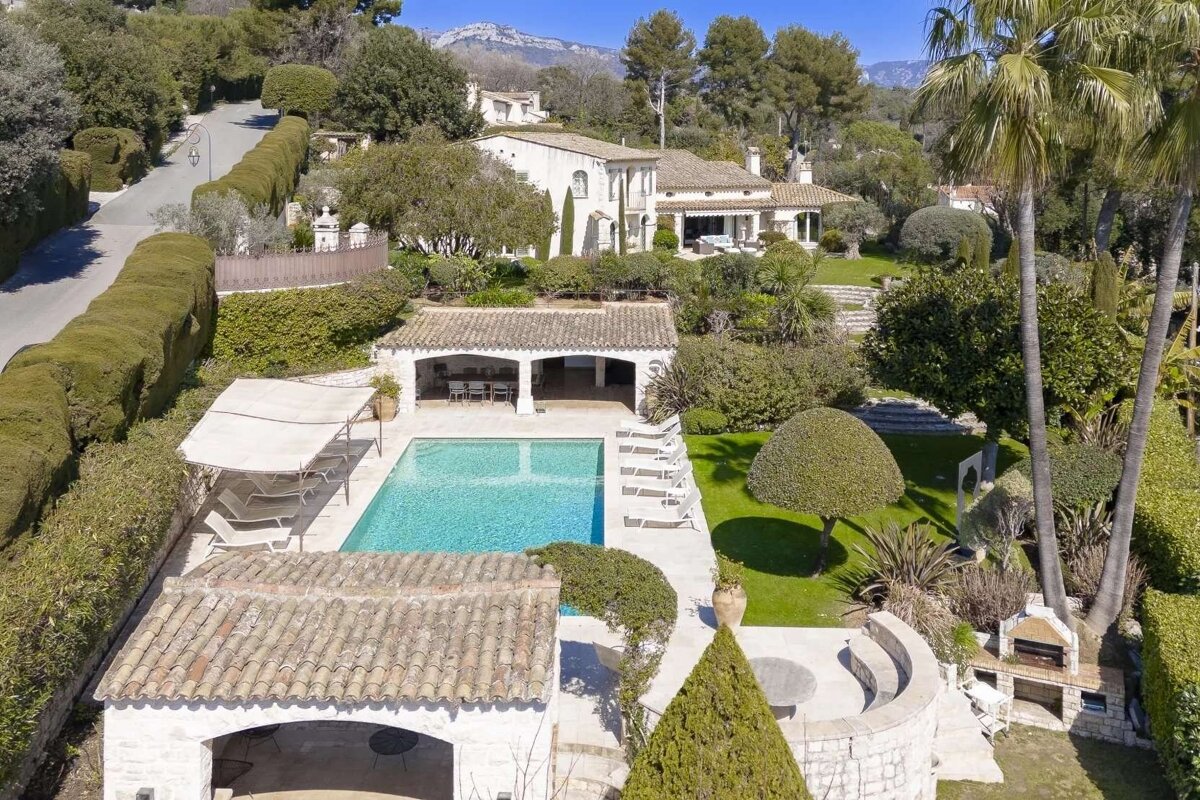 An expansive estate featuring a large white house, sparkling swimming pool, lush terraced gardens, and palm trees, set against a mountainous backdrop.