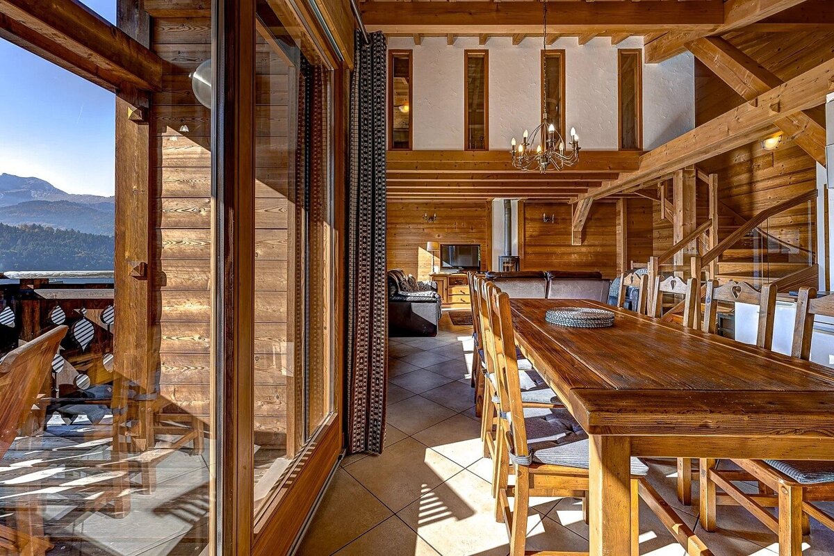Rustic wooden chalet interior featuring a long dining table and glass sliding doors that open to a scenic mountain balcony view.