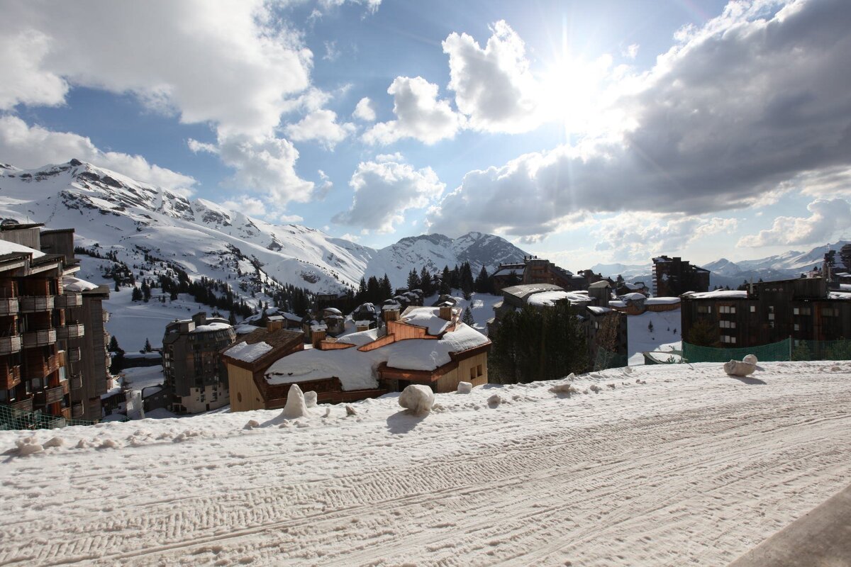 Sunny winter view of a snowy mountain village with alpine buildings, vast peaks, and bright clouds in the sky.