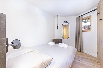 A minimalist modern bedroom with a white bed, mirror, wooden floor, and a window revealing a snowy mountain village. Clean design.