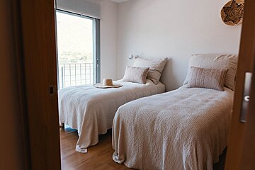 A bright, minimalist bedroom with two twin beds, neutral bedding, a straw hat, and a balcony overlooking a scenic water view.