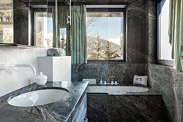 A luxurious dark marble bathroom with a sink, bathtub, green curtains, and a window revealing a snowy mountain view. A 'N°5' pillow rests by the tub.