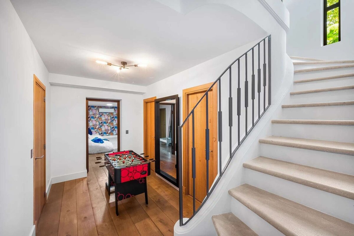 A white hallway with hardwood floors features a red foosball table, several wooden doors, and a curved staircase with a black metal railing.