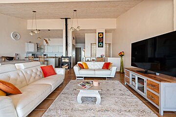 Bright, open-plan living room with white sofas, colorful cushions, large TV, wood stove, kitchen in background, and a soft rug on tiled floor.