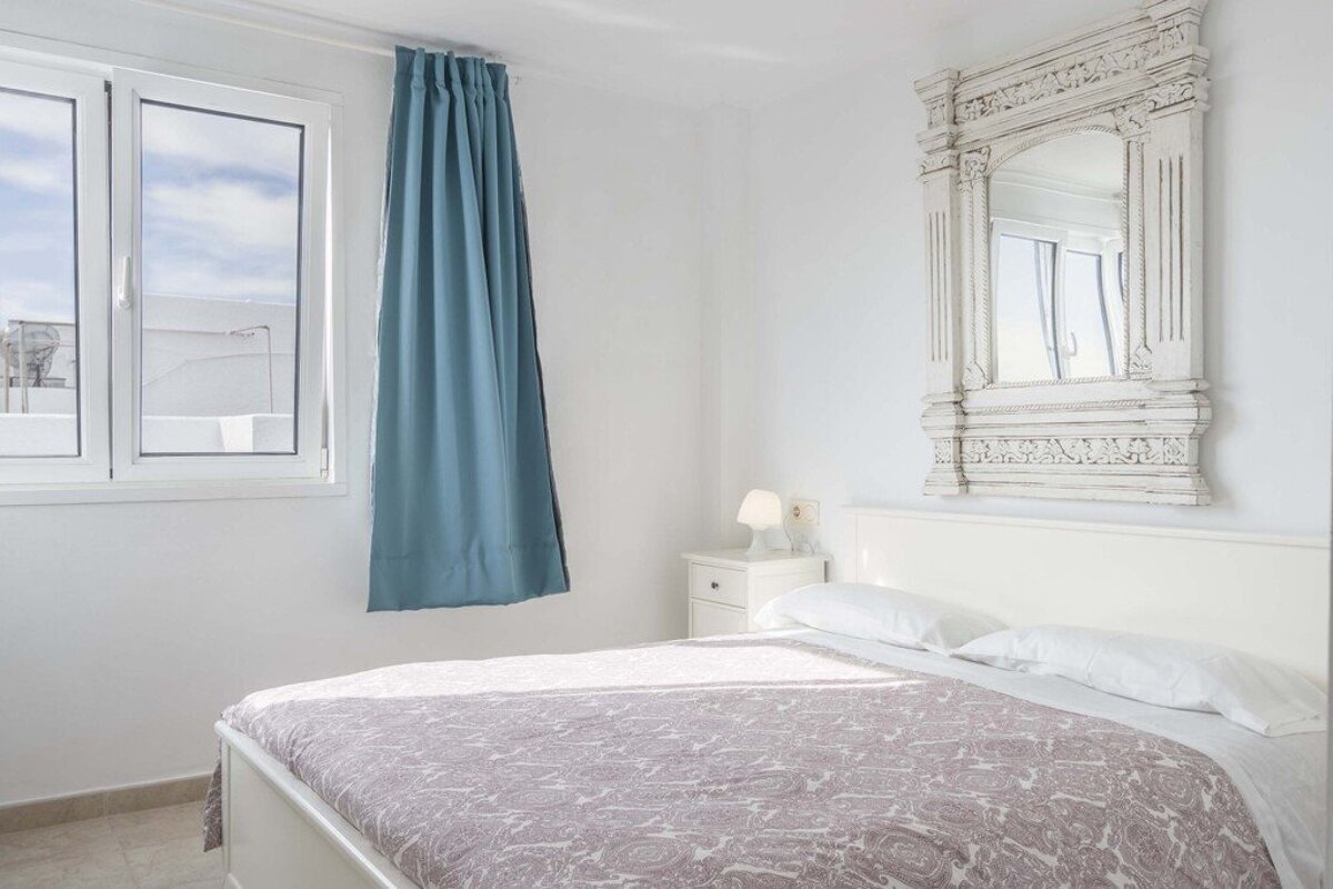 Bright white bedroom featuring a double bed, an ornate framed mirror above the headboard, and a window with a blue curtain.