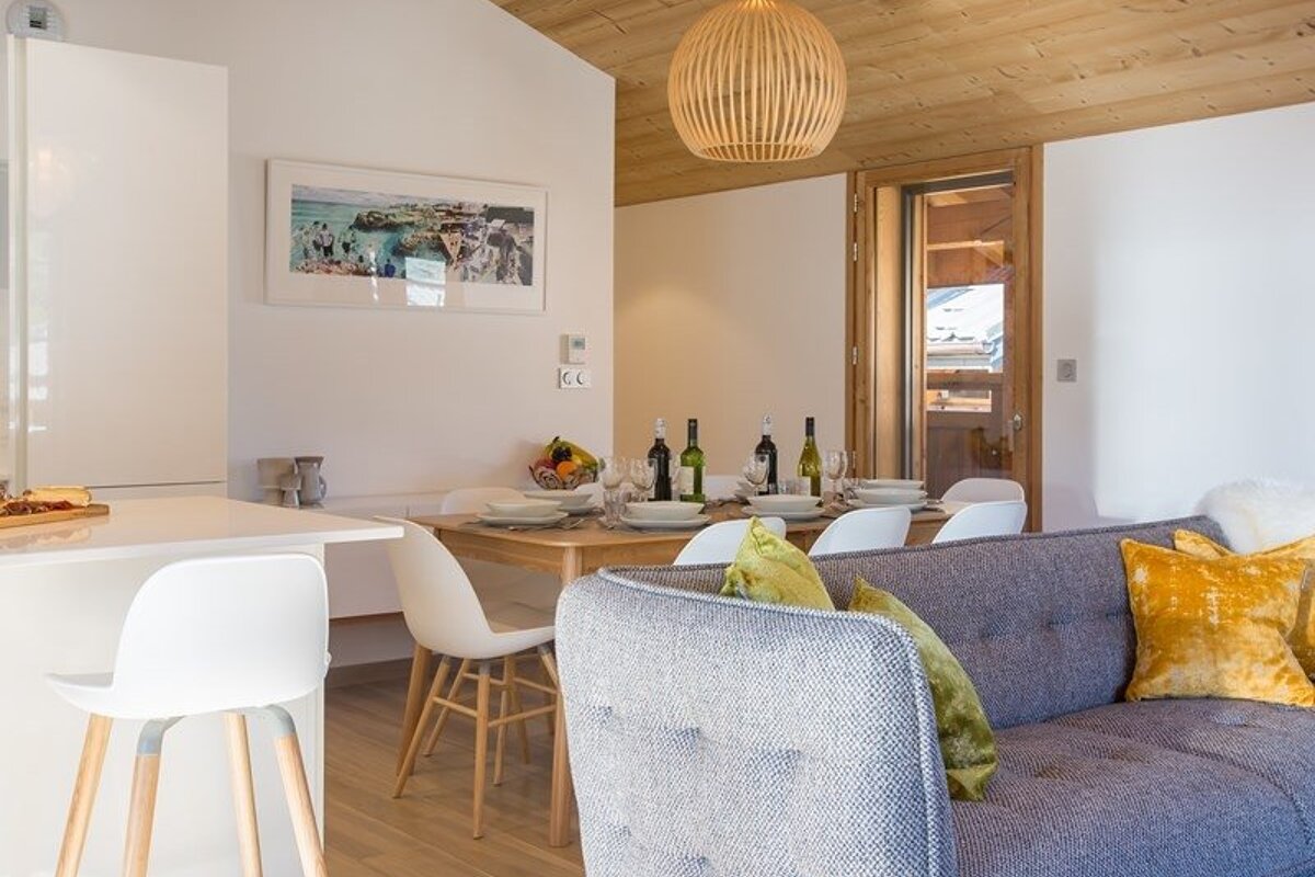 Modern open-plan living and dining room with a blue sofa, set wooden table, white chairs, and a warm wooden ceiling.